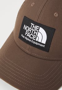 Brun hatt med en svart rektangulær lapp med hvit tekst og logo. Brodert tekstur og bøyd skjerm forbedrer designdetaljene.