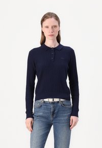 Pólo de malha canelada de cor azul-marinho com três botões e um pequeno logotipo. Combinado com jeans de lavagens claras e um cinto branco.