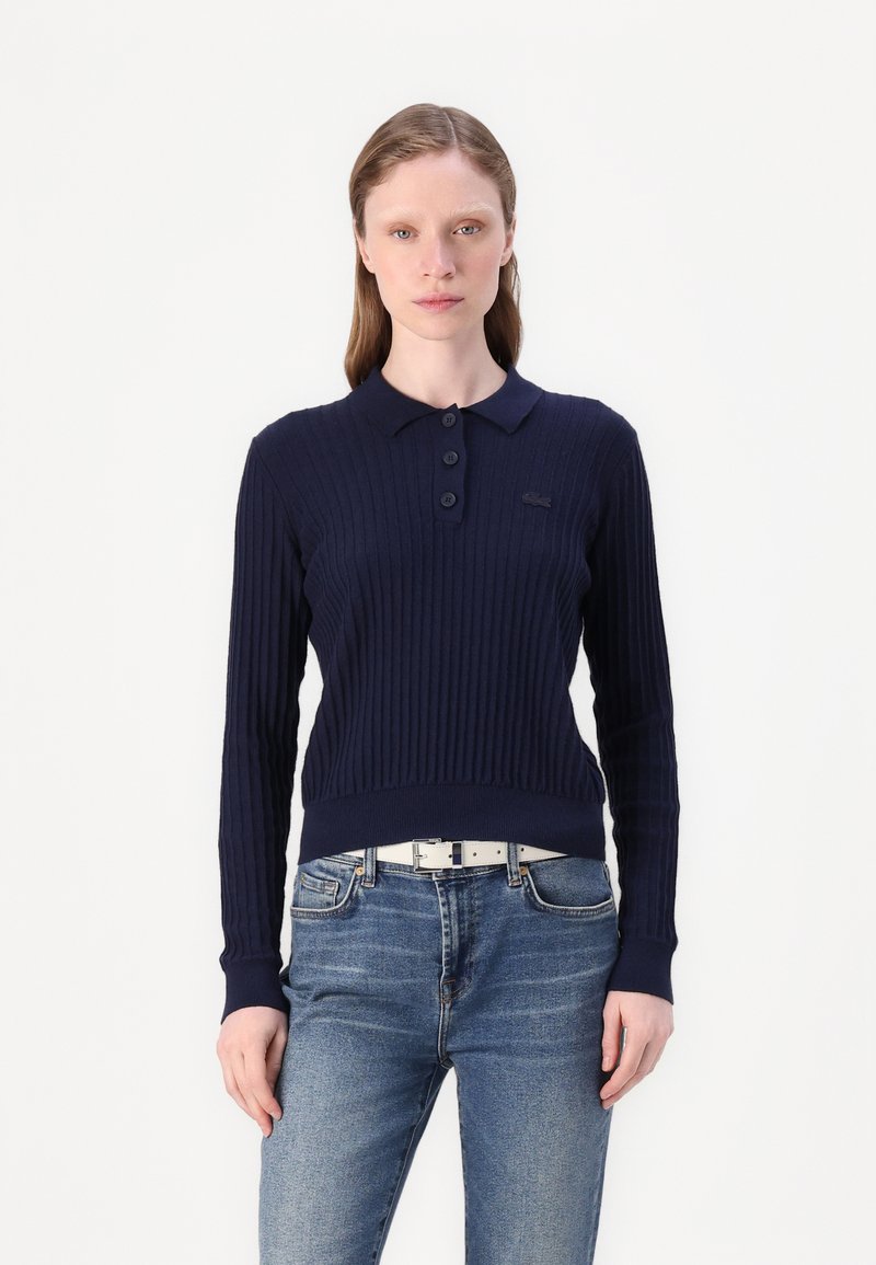 Pólo de malha canelada de cor azul-marinho com três botões e um pequeno logotipo. Combinado com jeans de lavagens claras e um cinto branco.