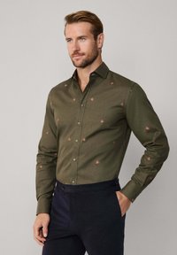 Man in een olijfgroene button-up shirt met kleine rode vogelprints en marineblauwe broek, staand met één hand in zijn zak.