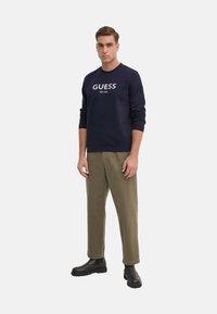 Felpa navy con logo "GUESS" sul davanti, abbinata a pantaloni verde oliva e stivali neri alla caviglia. Tessuto morbido e vestibilità comoda.