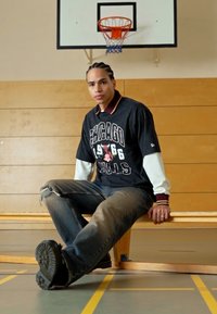 Črno oversize majica Chicago Bulls z belimi rokavi, unikatne modre kavbojke in črne športne copate. V ozadju lesena klop in košarkaški obroč.