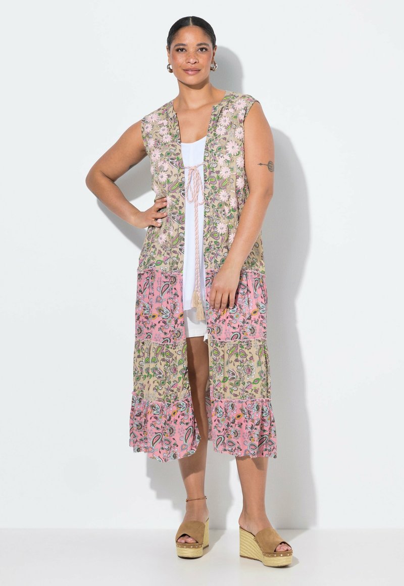 Gilet long à motif floral avec manches courtes, sections rose pastel et verte, ouvert à l'avant et avec un lien ajustable. Porté par-dessus une robe blanche.