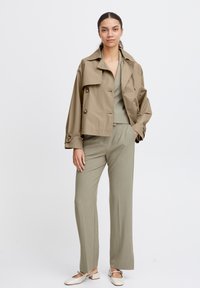 Cappotto trench corto kaki con bottoni doppi, abbinato a pantaloni larghi verdi oliva e ballerine crema. Tessuto morbido, design minimalista.