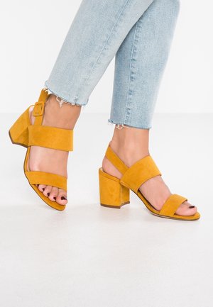 Fødder iført sennepsgule ruskindssandaler med blokhæl og ankelremme, kombineret med lyseblå cropped jeans med frynser.