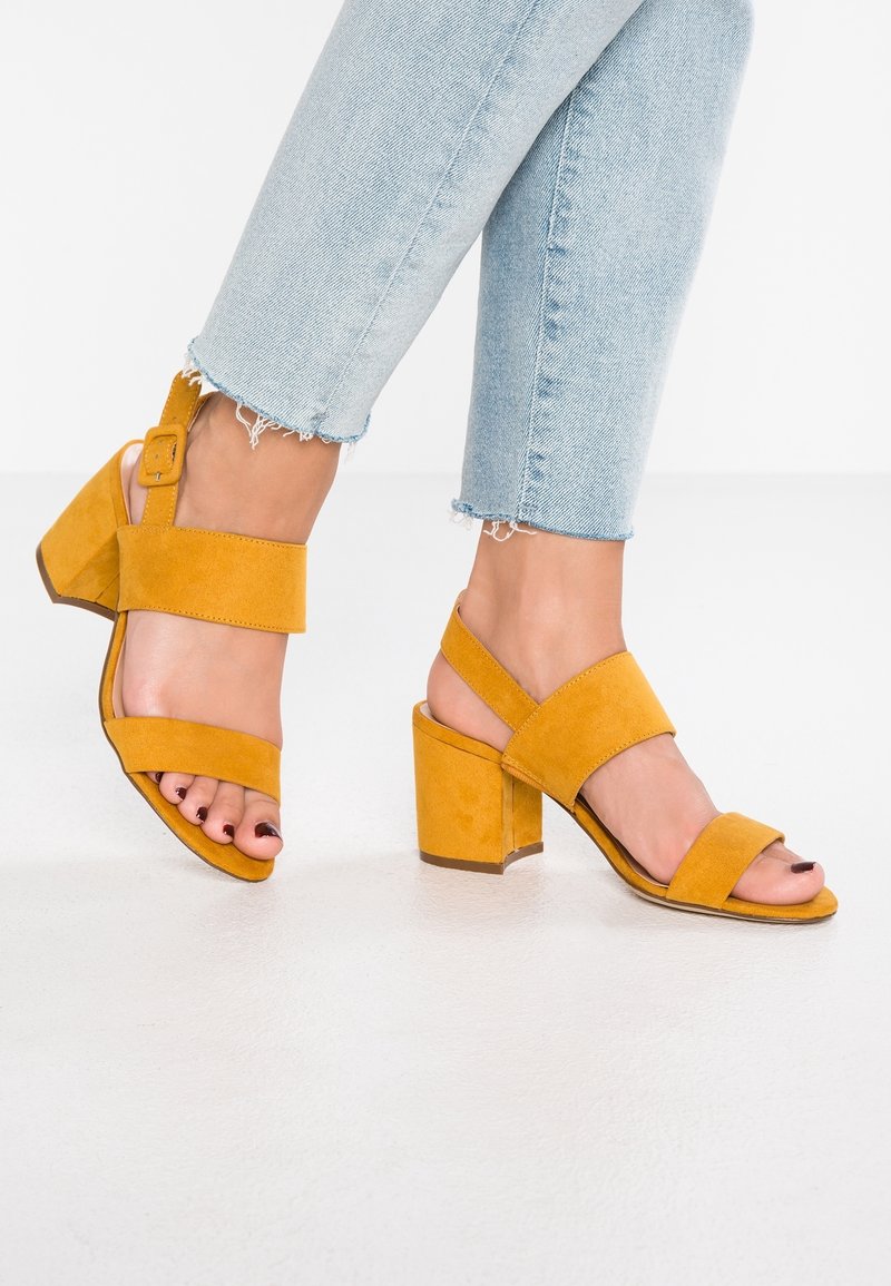 Pieds portant des sandales à talons blocs en daim jaune moutarde avec des brides à la cheville, assorties à un jean court bleu clair avec bords effilochés.