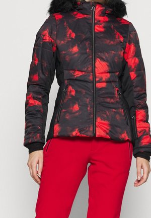 Veste zippée noire et rouge à motif avec capuche bordée de fourrure portée avec un pantalon rouge uni, montrée de la moitié du torse aux cuisses.