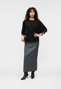 Μαύρο τοπ με κρόσσια και κοντά μανίκια, σε συνδυασμό με μια μακριά γκρι φούστα. Το μοντέλο φοράει μαύρα μυτερά μποτάκια μέχρι τον αστράγαλο και κρατά μια μικρή μαύρη τσάντα.