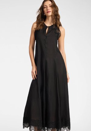 Kobieta w czarnej, bezrękawnej sukience maxi z zapięciem na guziki z przodu, z dekoltem typu keyhole, koronkową lamówką na ramionach i na dole, stojąca na białym tle.