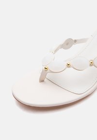 Λευκό επίπεδο σανδάλι με λουράκι Τ με οβάλ λευκές πέτρες και μικρές χρυσές τρουκς, κοντινή λήψη στην περιοχή των δαχτύλων και στις λεπτομέρειες του λουριού.