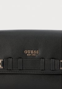 Svart strukturerad handväska med gyllene detaljer. Har "GUESS EST 1981"-logotyp och axelremmar. Slät yta med praktiska designelement.