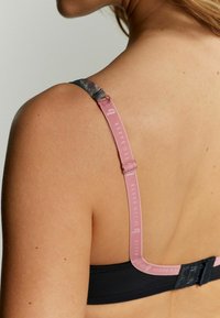 Zwarte beha met bloemenpatroon bandjes, voorzien van een verstelbare roze strap en een branded haak-en-oogsluiting aan de achterkant.