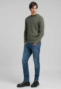 Maglione verde a trecce con scollo rotondo, abbinato a jeans blu e stivaletti neri alla caviglia. Il maglione presenta motivi a rilievo e polsini a coste.