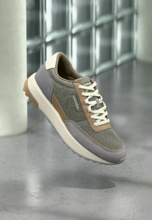 Codzienny sneaker z beżowymi, szarymi i siateczkowymi detalami, białymi sznurowadłami, beżową podeszwą, unoszący się nad wypolerowaną podłogą na tle ściany z szklanych cegieł.