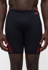 Torse et jambes masculins portant un jammers de natation Speedo noir avec des détails en zigzag rouges sur les côtés, debout, les bras détendus le long du corps.