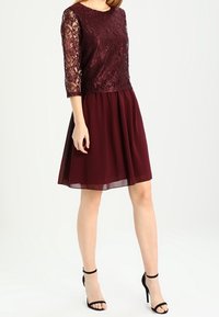 Burgundy klänning med spetsliv, trekvartsärmar och en flödande, genomskinlig kjol. Stylad med svarta klackar med remmar.