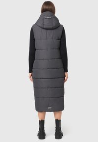 Pikk hall puffer vest koos kapuutsiga, millel on tikand, ja logo allservas, koos mustade pikkade saapatega.