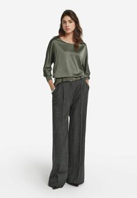Olivgrön satinblus med rundad halsringning, kombinerad med grå, högmidjade byxor med vida ben och mönster. Modellen står med händerna i fickorna.