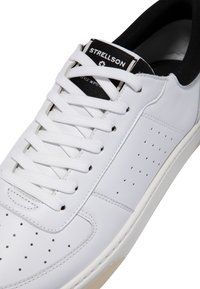 Sneaker bianco con tomaia in pelle liscia, lacci piatti, dettagli perforati sul lato e imbottitura interna nera. Presenta un'etichetta con il marchio.