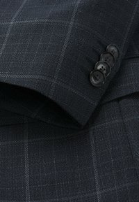 Tessuto strutturato blu navy con un sottile motivo a quadri, caratterizzato da quattro bottoni neri sul polsino, che evidenziano elementi di design sartoriale.