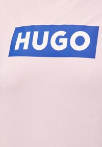 Άσπρο ύφασμα με τη λέξη «HUGO» με έντονα λευκά γράμματα μέσα σε ένα συμπαγές μπλε ορθογώνιο φόντο.