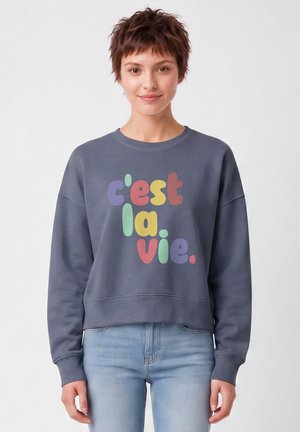 Jeune femme aux cheveux courts portant un sweat gris avec un texte coloré "c'est la vie" et un jean bleu clair, debout devant un fond uni.