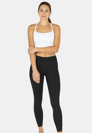 Junge Frau mit langem, zurückgebundenem braunem Haar, trägt einen weißen Sport-BH und schwarze Leggings, steht mit einem verschränkten Arm.