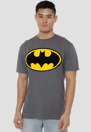 Young man wearing a gray t-shirt with a large yellow and black Batman logo, standing against a plain light background.