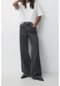 Leveälahkeiset farkut haalistuneesta mustasta denimistä, korkeavyötäröiset ja hieman teksturoidut, yhdistettynä löysään valkoiseen t-paitaan ja mustiin, teräväkärkiisiin kenkiin.