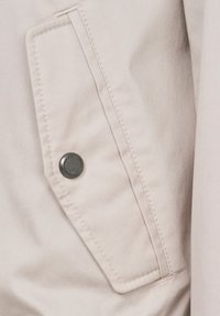 Beige Jackendetail mit einer großen Klappentasche, einem runden silbernen Knopf und kontrastierenden weißen Nähten auf einem weichen Stoff.