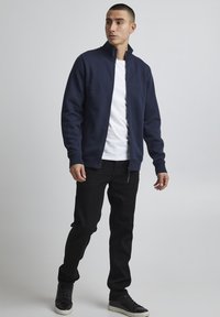 Marineblå zip-up jakke med høy krave, over en hvit t-skjorte, parret med svarte jeans og svarte sneakers, med oppbrettede ermer og en jevn tekstur.