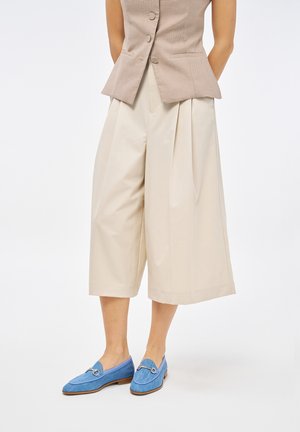 Culotte beige plissettate con tasche, abbinate a scarpe blu slip-on con parte superiore texturizzata e dettaglio in metallo sulla tomaia.
