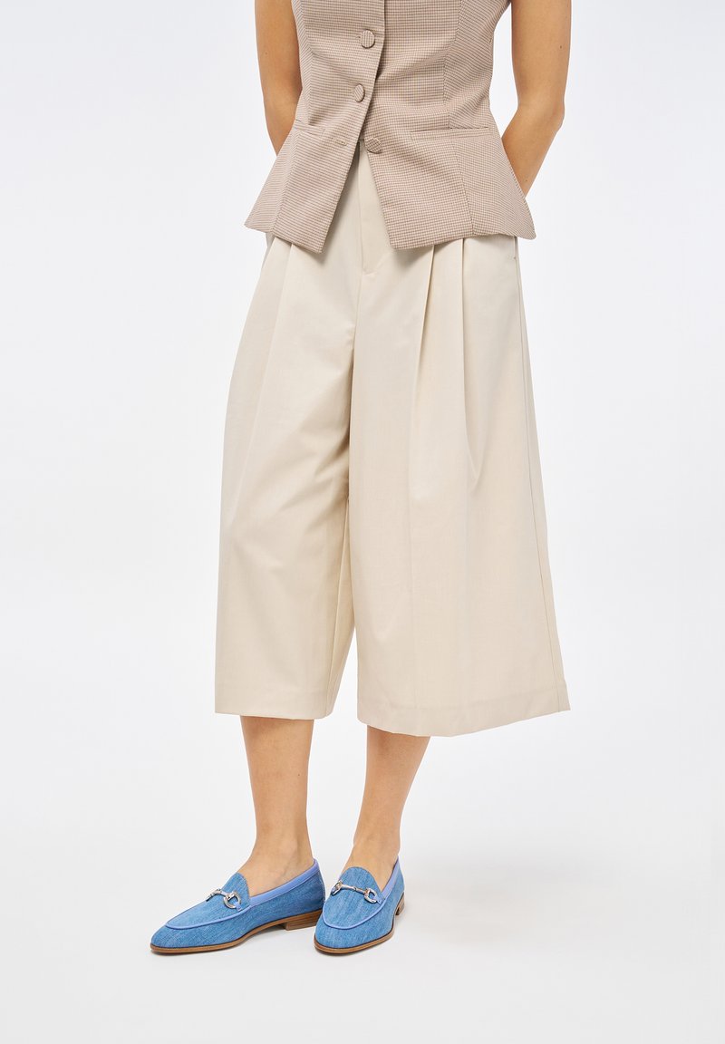 Culotte beige plissettate con tasche, abbinate a scarpe blu slip-on con parte superiore texturizzata e dettaglio in metallo sulla tomaia.