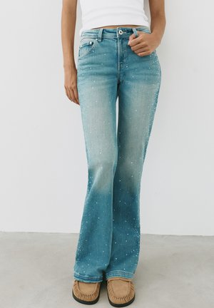 Femme portant un jean taille haute bleu clair avec des taches blanches, un haut blanc et des mocassins couleur beige, debout devant un fond blanc.