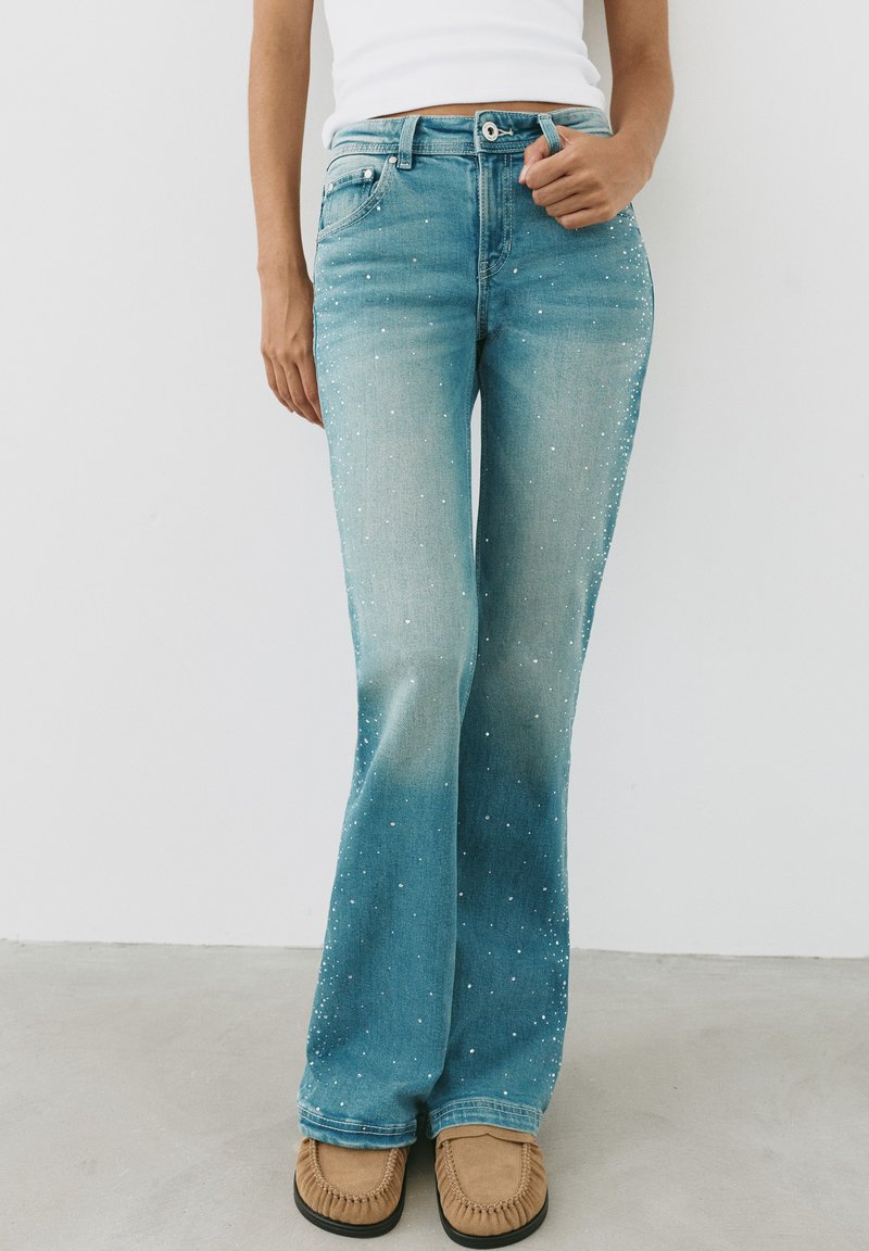 Femme portant un jean taille haute bleu clair avec des taches blanches, un haut blanc et des mocassins couleur beige, debout devant un fond blanc.