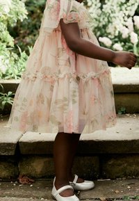 Robe rose pâle avec un imprimé floral, volants superposés et tissu transparent. Assortie à des chaussures Mary Jane argentées sur les pieds d'un enfant.