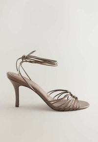 Sandale marron à talons hauts avec de fines brides traversant le pied et un lien à nouer à la cheville sur un fond clair uni.