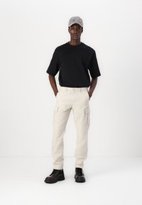 Czarny t-shirt krótki rękaw, beżowe spodnie cargo i czarne buty sznurowane. Szara czapka z ozdobami. Model stoi z rękami w kieszeniach.