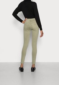 Jeans skinny de tiro alto en color verde oliva, con bolsillos traseros, textura suave y pierna ajustada. Combinados con una blusa negra de manga larga y zapatos negros.