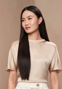 Femme aux longs cheveux lisses et foncés, portant un chemisier en satin beige et un pantalon blanc, regardant légèrement sur le côté avec un fond neutre.