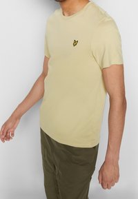 Jasnożółta bawełniana koszulka z okrągłym dekoltem, krótkimi rękawami oraz małym żółtym haftowanym logo ptaka na piersi.
