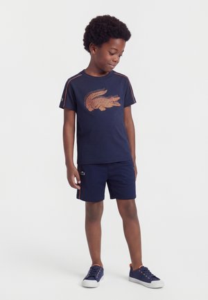 Ragazzo con maglietta blu navy con una grande grafica di coccodrillo arancione, pantaloncini abbinati e scarpe da ginnastica blu navy con suola bianca, in piedi con lo sguardo rivolto verso il basso.