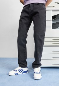 Svarte denimbukser med rett snitt, sammen med hvite joggesko med blå detaljer. Bakgrunnen viser en hvit skriver. Blå teppe under.