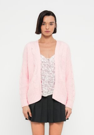 Ροζ ανοιχτό ροζ πλεκτό ζακέτα με σχέδιο πλεξίδας, ανοιχτό μπροστινό μέρος και μακριά μανίκια, φορεμένο πάνω από ένα φλοράλ μπλουζάκι και μαύρη πλισέ φούστα.