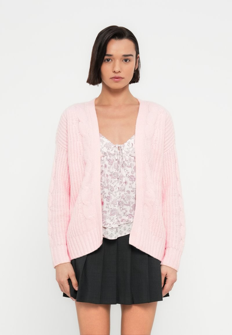 Ροζ ανοιχτό ροζ πλεκτό ζακέτα με σχέδιο πλεξίδας, ανοιχτό μπροστινό μέρος και μακριά μανίκια, φορεμένο πάνω από ένα φλοράλ μπλουζάκι και μαύρη πλισέ φούστα.