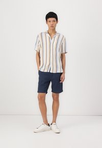 Camisa de manga corta con botones, de rayas verticales en beige y azul marino; combinada con pantalones cortos azul marino y zapatillas blancas. Ajuste relajado, diseño casual.