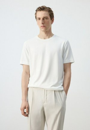 Hombre joven con cabello corto y marrón, vistiendo una camiseta blanca lisa y pantalones beige claros, de pie con una mano en el bolsillo contra un fondo blanco.