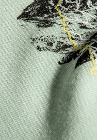 Tekstilstoff i lys mintgrønn med svart og gul broderitdesign. Synlig tekstur med diagonalt vevemønster, litt rynket.