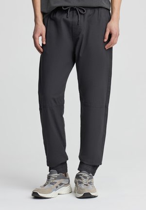 Persona che indossa pantaloni jogger neri con polsini elastici e scarpe sportive grigie, beige e bianche, in piedi su uno sfondo grigio chiaro.