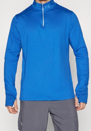 Homme portant un maillot de sport bleu à manches longues avec fermeture éclair au quart et un short gris, debout les mains détendues le long du corps.